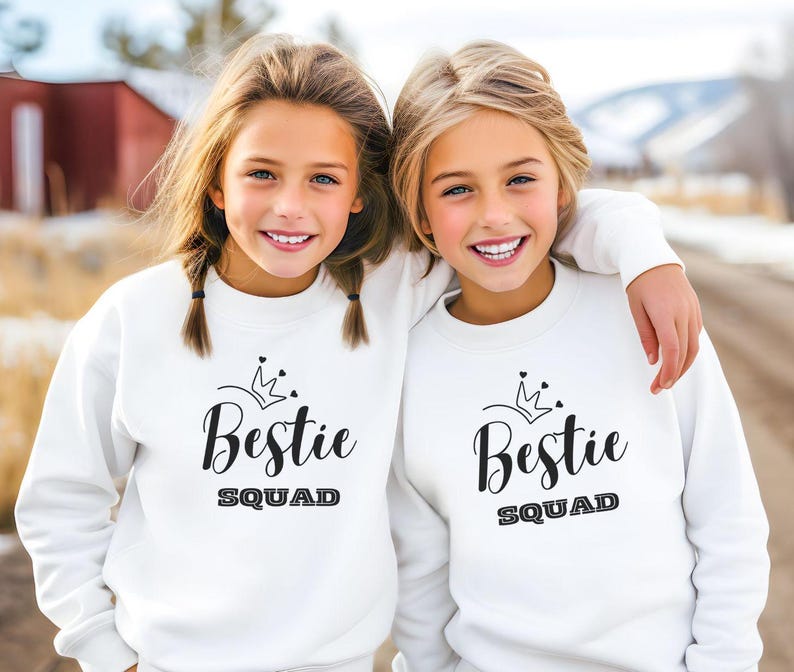 Puede incluir: Dos chicas con sudaderas blancas con el texto "Bestie Squad" y un gr&aacute;fico de corona. Las chicas sonr&iacute;en y miran a la c&aacute;mara.
