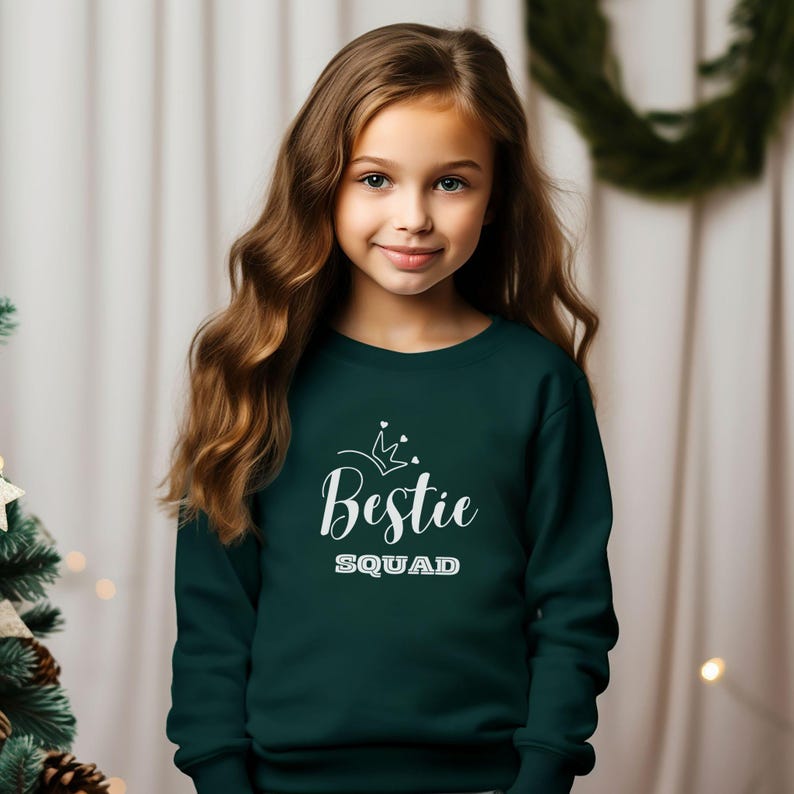Puede incluir: Una sudadera verde oscuro con un gr&aacute;fico blanco que dice "Bestie Squad" con una corona y corazones sobre el texto.