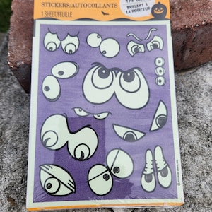 May include: A sheet of Hallmark glow-in-the-dark stickers featuring various cartoon eye designs on a purple background. The packaging includes the text "STICKERS/AUTOCOLLANTS", "GLOW IN THE DARK", and "BRILLANT À LA NOIRCEUR" with small bat and pumpkin icons.