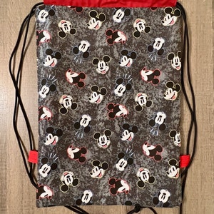 Puede incluir: Una bolsa de cord&oacute;n negro con la parte superior roja y un estampado de Mickey Mouse. La bolsa tiene cordones negros y la parte superior roja.