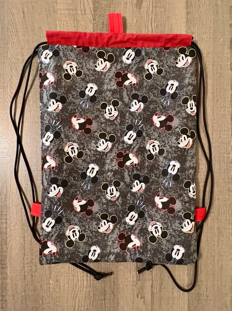 Puede incluir: Una bolsa de cord&oacute;n negro con la parte superior roja y un estampado de Mickey Mouse. La bolsa tiene cordones negros y la parte superior roja.