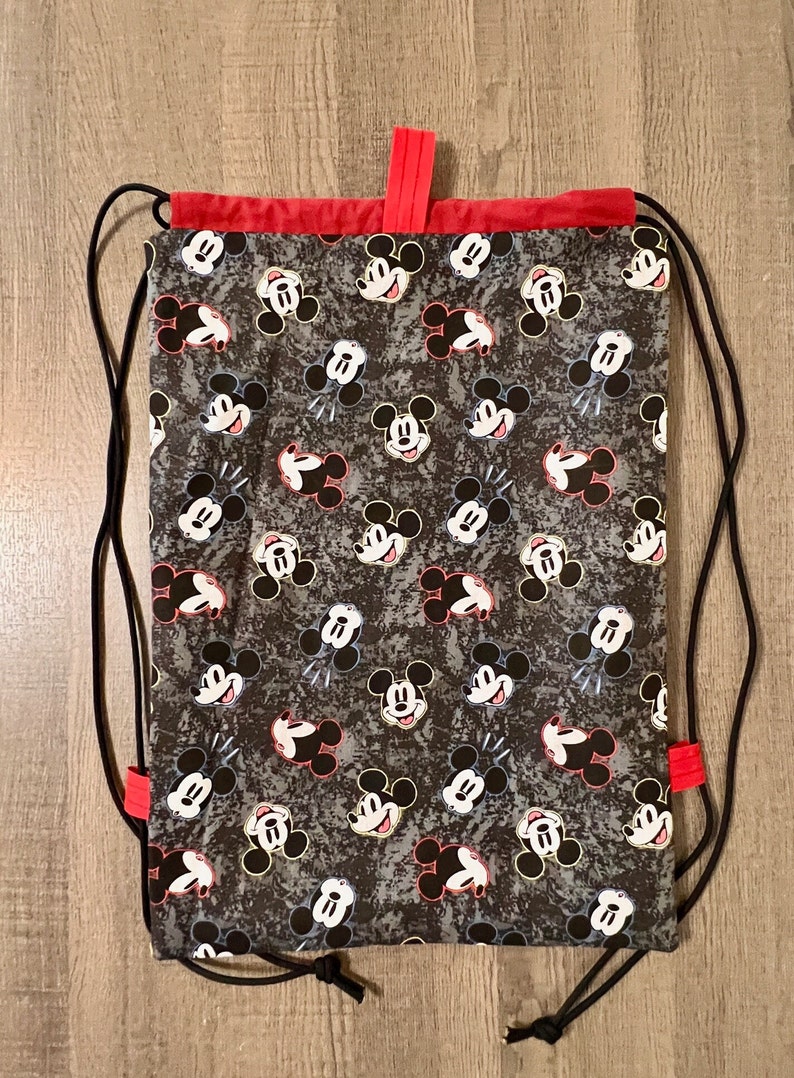 Puede incluir: Una bolsa de cord&oacute;n negro con ribete rojo y estampado de Mickey Mouse. La bolsa tiene un cierre de cord&oacute;n negro y est&aacute; hecha de un tejido duradero.