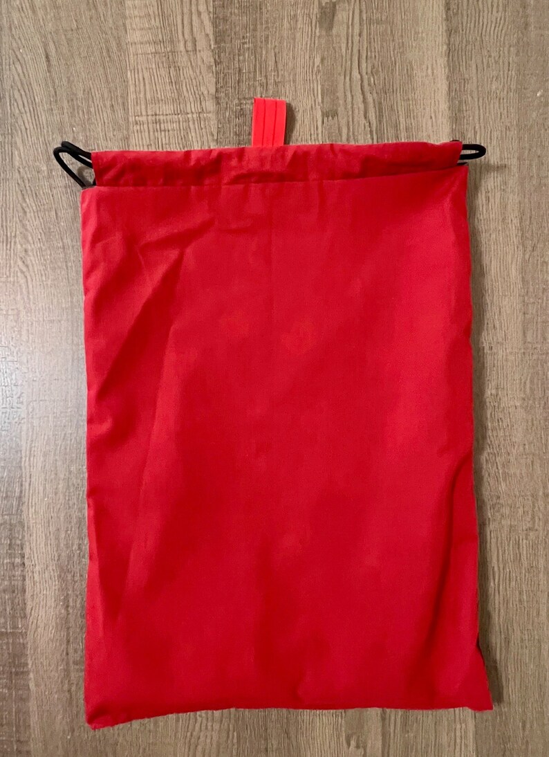 Puede incluir: Una bolsa de cord&oacute;n roja con un cord&oacute;n negro y una leng&uuml;eta roja.