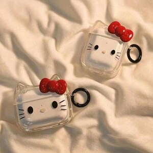 May include: Two clear AirPod cases with a Hello Kitty design. Each case has a white face with black eyes, whiskers, and a red bow. The cases have a black ring for attachment.