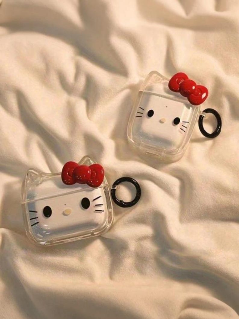 May include: Two clear AirPod cases with a Hello Kitty design. Each case has a white face with black eyes, whiskers, and a red bow. The cases have a black ring for attachment.