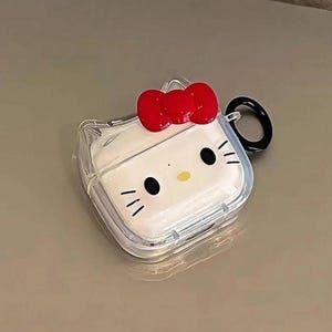 May include: Clear protective case for wireless earbuds, featuring a white face design with black eyes, whiskers, and a yellow nose. A red bow sits atop the case, and a black ring is attached for portability. The case is designed to resemble a popular cartoon character.