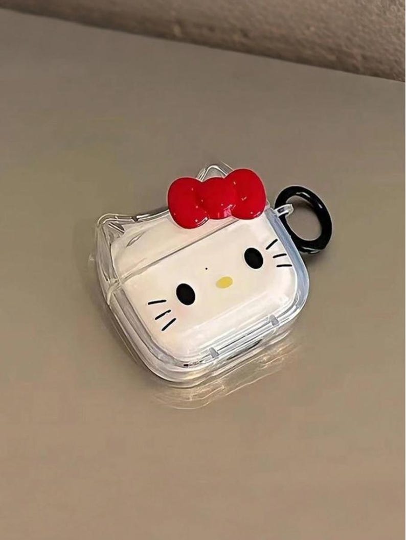 May include: Clear protective case for wireless earbuds featuring a white design with a cartoon face, black eyes, and whiskers. A red bow is at the top. The case has a black ring for attachment.
