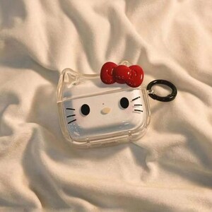 May include: Clear, transparent AirPods case with a red bow and a cartoon face design. The case has a black ring for attachment. The design is a popular character. The case is on a white, textured surface.