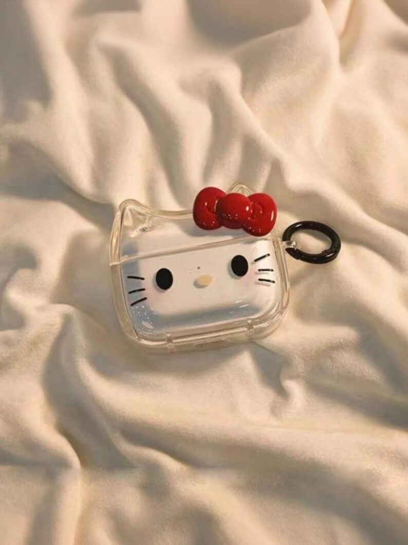 May include: Clear, transparent AirPods case with a red bow and a cartoon face design. The case has a black ring for attachment. The design is a popular character. The case is on a white, textured surface.