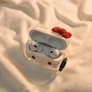 May include: Clear protective case for wireless earbuds, featuring a Hello Kitty design with a red bow on top. The case is open, revealing white earbuds inside. A black carabiner clip is attached to the side.