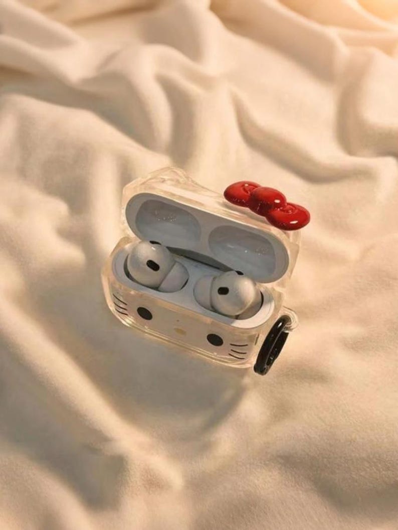 May include: Clear protective case for wireless earbuds, featuring a Hello Kitty design with a red bow on top. The case is open, revealing white earbuds inside. A black carabiner clip is attached to the side.