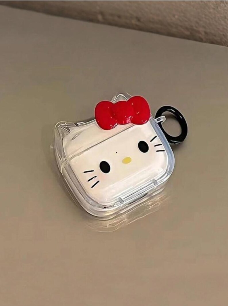 May include: Clear AirPod case featuring a white face with black eyes, whiskers, and a yellow nose, topped with a red bow. The case has a black ring for attachment.
