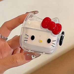 May include: Clear, square-shaped AirPod case featuring a white Hello Kitty design with black eyes, whiskers, and a yellow nose. A red bow adorns the top. The case is held by a hand.