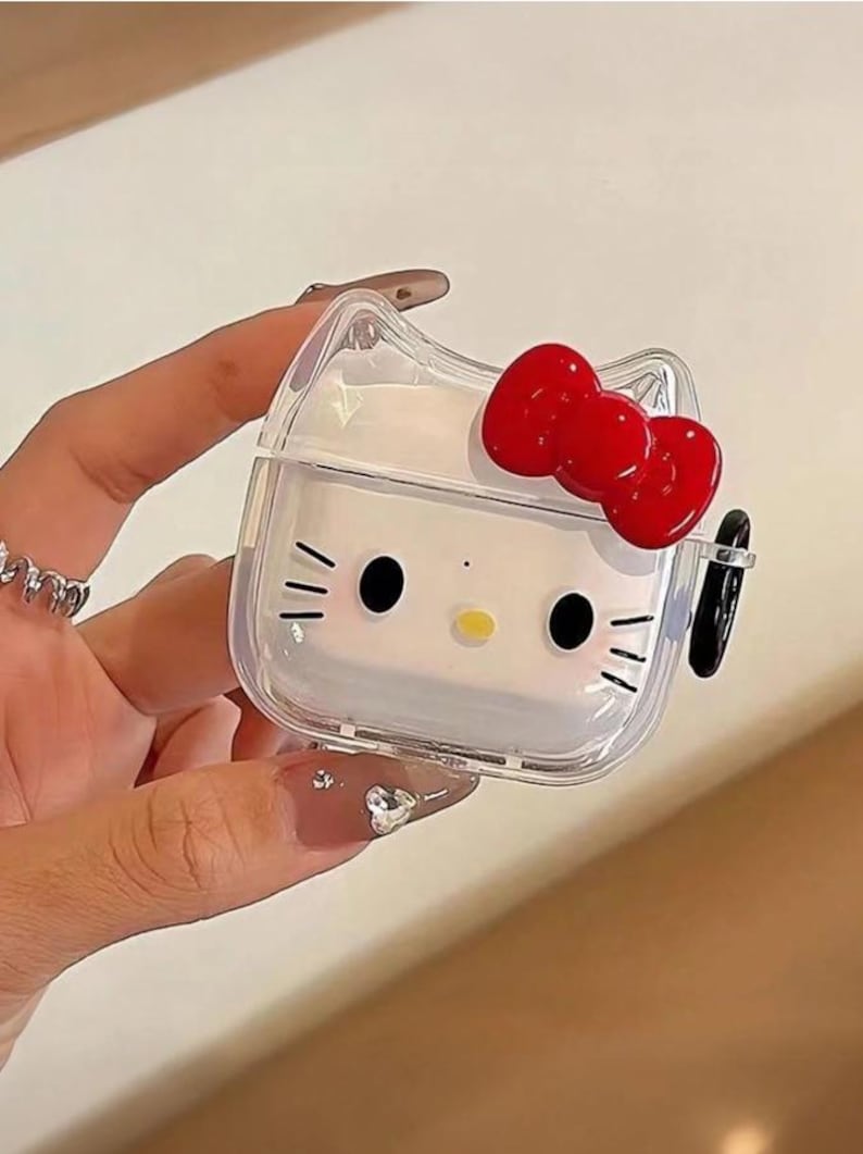 May include: Clear, square-shaped AirPod case featuring a white Hello Kitty design with black eyes, whiskers, and a yellow nose. A red bow adorns the top. The case is held by a hand.