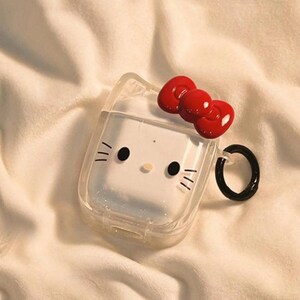 May include: Clear AirPod case featuring a white cartoon cat face with black eyes, whiskers, and a red bow. The case has a black ring for attachment and is set against a soft, white background.
