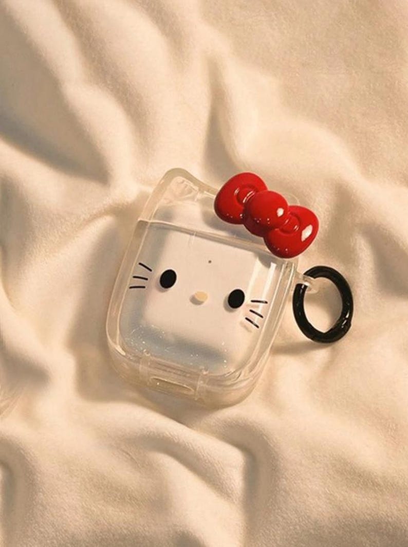 May include: Clear AirPod case featuring a white cartoon cat face with black eyes, whiskers, and a red bow. The case has a black ring for attachment and is set against a soft, white background.
