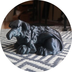 Peut inclure: Sculpture noire stylisée d'un mouton en position de repos. La sculpture a une surface texturée et est placée sur un fond à motifs. Le mouton a un long nez et est couché.