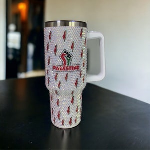 Op de afbeelding: Witte reistumbler van roestvrij staal met een witte handgreep. De tumbler is bedekt met strass en heeft een rode, groene en zwarte vuist met het woord "PALESTINE" in rode letters.