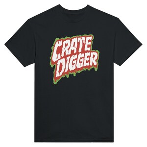 May include: A black short-sleeve t-shirt featuring a graphic on the chest with the text CRATE DIGGER in white letters. The text has a red outline and a green dripping effect underneath, creating a bold, high-contrast design on the dark fabric.