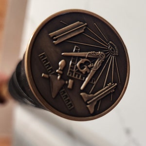 Puede incluir: Un emblema masónico de metal de color bronce con un compás y una escuadra, un nivel, una paleta y un ojo que todo lo ve.