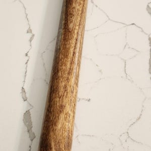 May include: A wooden-handled tool with a black top and gold-colored accents. The handle has a wood grain pattern. The tool is likely used for a specific purpose, possibly related to smoking or herbs.