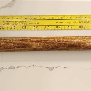 May include: A wooden pool cue with a black tip and a yellow ruler. The cue is approximately 57 inches long. The cue has a light brown wood grain pattern. The ruler is yellow with black markings.
