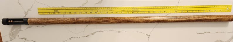 May include: A wooden pool cue with a black tip and a yellow ruler. The cue is approximately 57 inches long. The cue has a light brown wood grain pattern. The ruler is yellow with black markings.