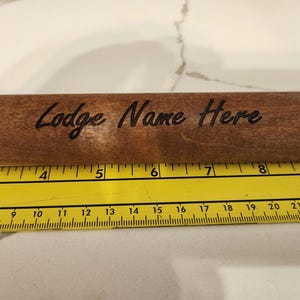 May include: A wooden rolling pin with the text "Lodge Name Here" engraved on its surface. The rolling pin is brown and is placed next to a yellow ruler. The ruler shows measurements in inches.
