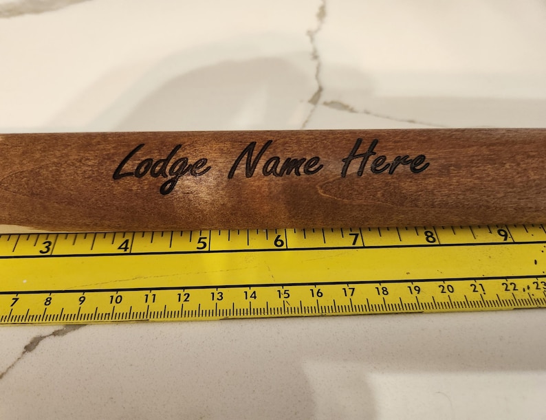 May include: A wooden rolling pin with the text "Lodge Name Here" engraved on its surface. The rolling pin is brown and is placed next to a yellow ruler. The ruler shows measurements in inches.