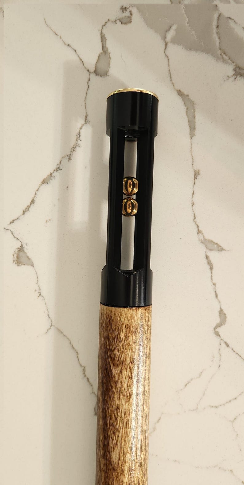 May include: A black and gold cue stick with a wooden handle. The top of the cue stick is black with a gold band. The middle section is black with gold accents. The bottom is a light brown wood.