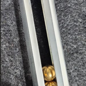 May include: A silver-colored metal object with a rectangular opening revealing two gold-colored spheres. The top has a gold-colored band. The object is on a dark gray surface.