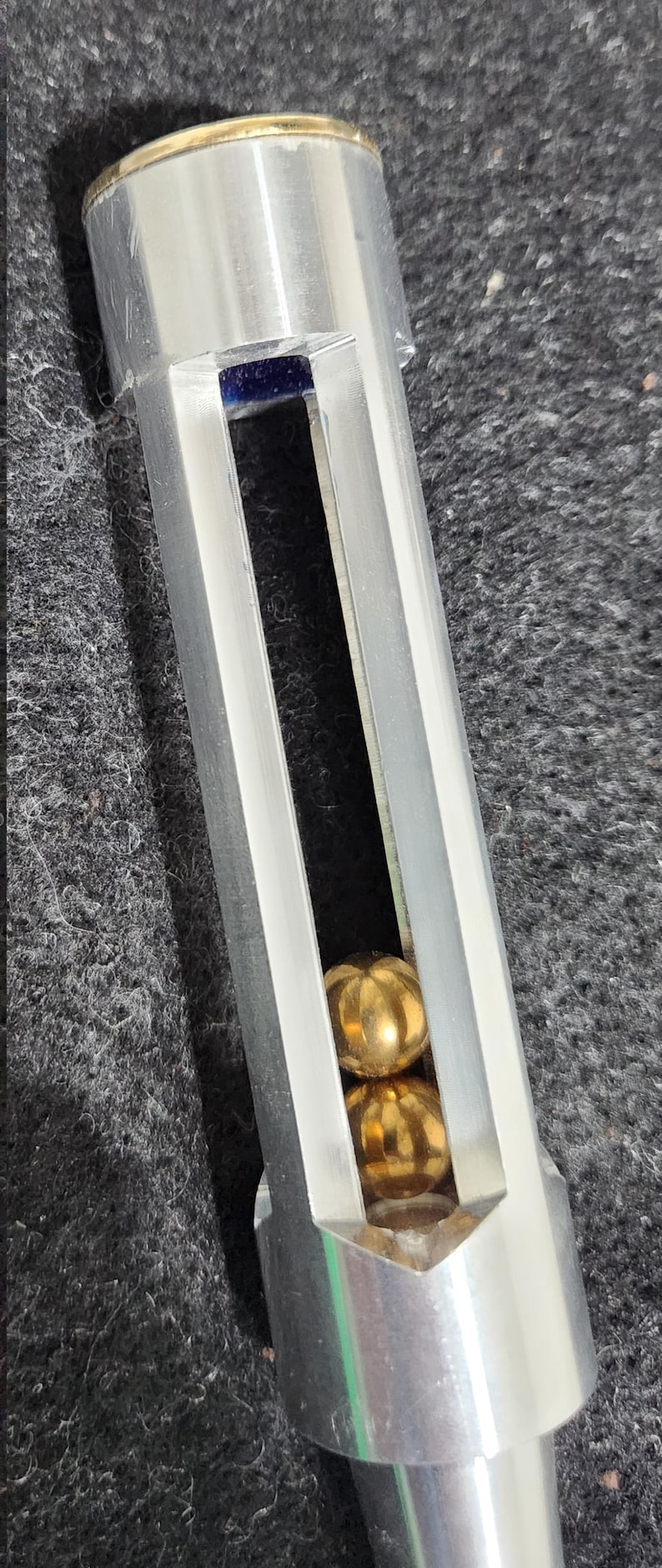 May include: A silver-colored metal object with a rectangular opening revealing two gold-colored spheres. The top has a gold-colored band. The object is on a dark gray surface.