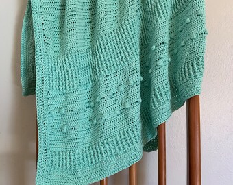 May include: A light green crocheted baby blanket with a textured pattern. The blanket is draped over a wooden chair.