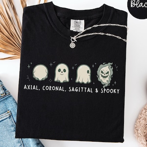 Peut inclure: Un t-shirt noir avec un motif représentant quatre illustrations de fantômes et le texte "AXIAL, CORONAL, SAGITTAL & SPOOKY" imprimé en blanc. Le design comprend une forme de crâne circulaire, un fantôme simple, un fantôme de profil et un fantôme effrayant.