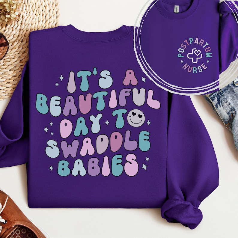 May include: Purple sweatshirt with the text "It's a beautiful day to swaddle babies" in colorful, retro-style lettering. The sweatshirt also has a small, white patch with the text "Postpartum Nurse" and a cross.