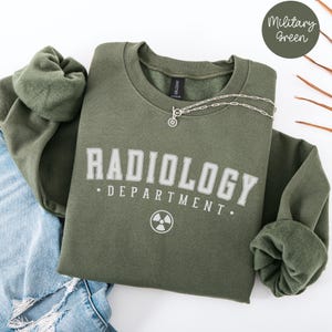Könnte beinhalten: Ein militärgrüner Pullover mit dem Text "RADIOLOGY DEPARTMENT." in weißen Buchstaben und einem weißen Strahlungssymbol.