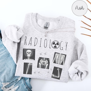 Könnte beinhalten: Ein graues Sweatshirt mit dem Text "RADIOLOGY" und einem Strahlungssymbol. Das Sweatshirt zeigt mehrere Röntgenbilder von Knochen.