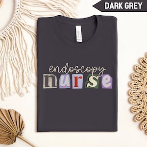 May include: Dark grey t-shirt with the text "endoscopy nurse" in colorful, cut-out letters.