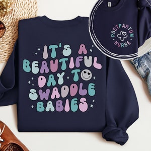 May include: Navy blue sweatshirt with the text "It's a beautiful day to swaddle babies" in colorful, retro-style lettering. The text includes a smiley face with hearts for eyes.