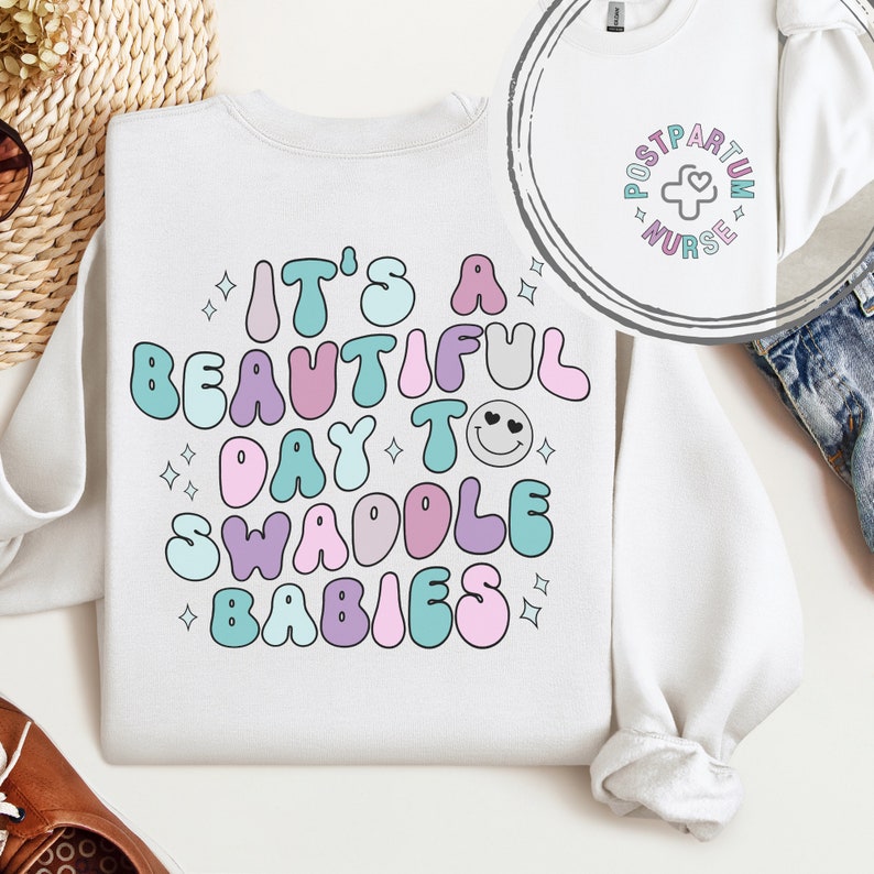 May include: White sweatshirt with a colorful graphic that says "It's a beautiful day to swaddle babies". The graphic features a smiley face with hearts for eyes and stars.