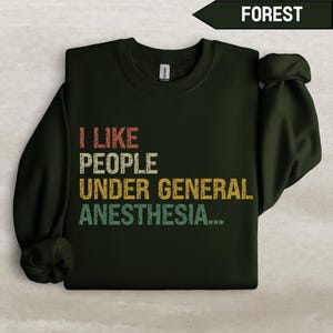 Könnte beinhalten: Ein dunkelgrüner Pullover mit dem Text "ICH MAG MENSCHEN UNTER ALLGEMEINER NARKOSE..." in einer Vintage-Schriftart mit bunten Akzenten.