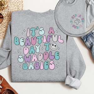 May include: A gray sweatshirt with the text "It's a beautiful day to swaddle babies" in colorful, retro-style lettering. The text is surrounded by small stars and a smiley face with hearts for eyes.
