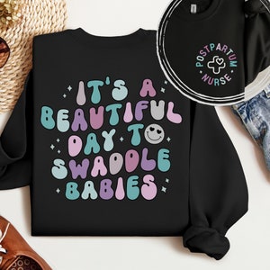 May include: Black sweatshirt with a colorful graphic that says "It's a beautiful day to swaddle babies". The graphic features a smiley face with hearts for eyes.