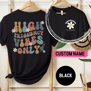 Op de afbeelding: Zwart T-shirt met een kleurrijke afbeelding met de tekst "High Frequency Vibes Only" en een klein beeld van een echografiekop. De achterkant van het shirt heeft een witte madeliefje met de tekst "Sonographer Elizabeth" in zwart.