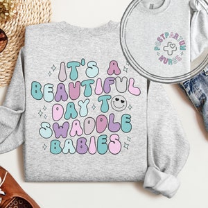 May include: A gray sweatshirt with the text "It's a beautiful day to swaddle babies" in colorful, bubble-like letters. The text is surrounded by small white stars.