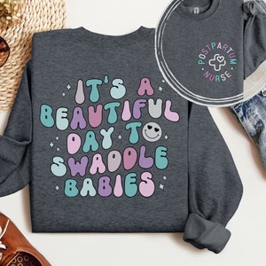 May include: A dark gray sweatshirt with the text "It's a beautiful day to swaddle babies" in colorful, retro-style lettering. The text is surrounded by small white stars.
