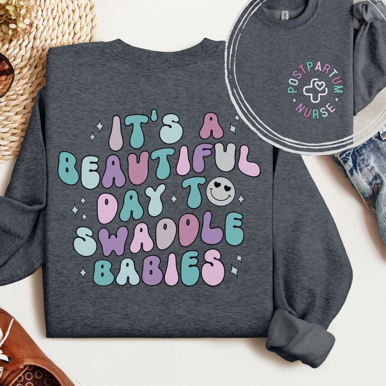 May include: A dark gray sweatshirt with the text "It's a beautiful day to swaddle babies" in colorful, retro-style lettering. The text is surrounded by small white stars.