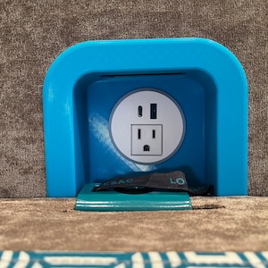 May include: A blue plastic outlet cover with a standard US electrical outlet and two USB ports. The cover is mounted on a brown fabric surface.