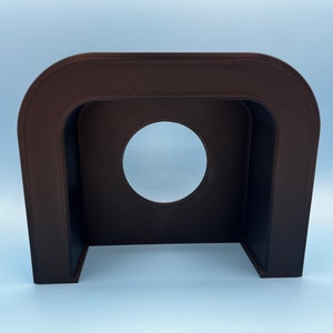 May include: A dark brown plastic 3D printed stand with a circular opening in the center. The stand is designed to hold a small object, such as a phone or a tablet.