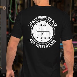 以下が含まれることがあります： 黒いTシャツに、白いスティックシフトギアのパターンが円形に描かれており、その周りに「車両は盗難防止装置付き」という文字が書かれています。